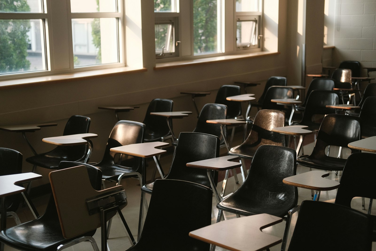 Rows of empty classroom chairs with fold-down desks in late-afternoon light from a bank of windows, the kind of room whose students and families a school holds records about.